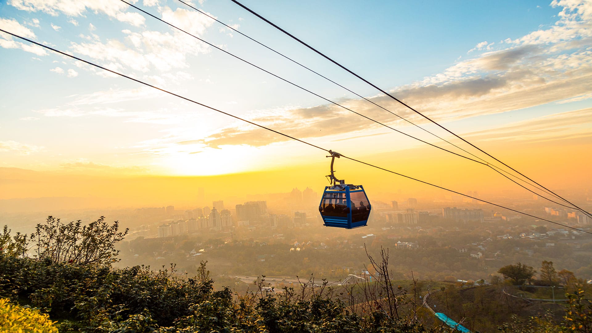 "A vibrant sunset view of Kok Tobe with a cable car gliding over the cityscape, surrounded by lush greenery and golden skies.