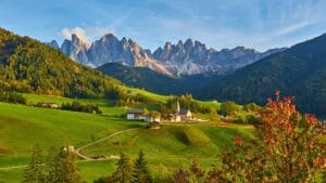 Scenic view of Bolzano with lush green meadows, scattered houses, and the Dolomites in the background under a clear sky.