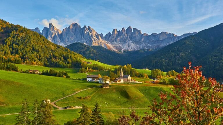 Scenic view of Bolzano with lush green meadows, scattered houses, and the Dolomites in the background under a clear sky.