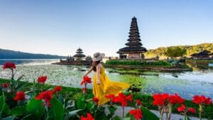 A vibrant scene of Ulun Danu Beratan Temple in Bali, with a woman in a flowing yellow dress surrounded by red flowers and serene lake waters.