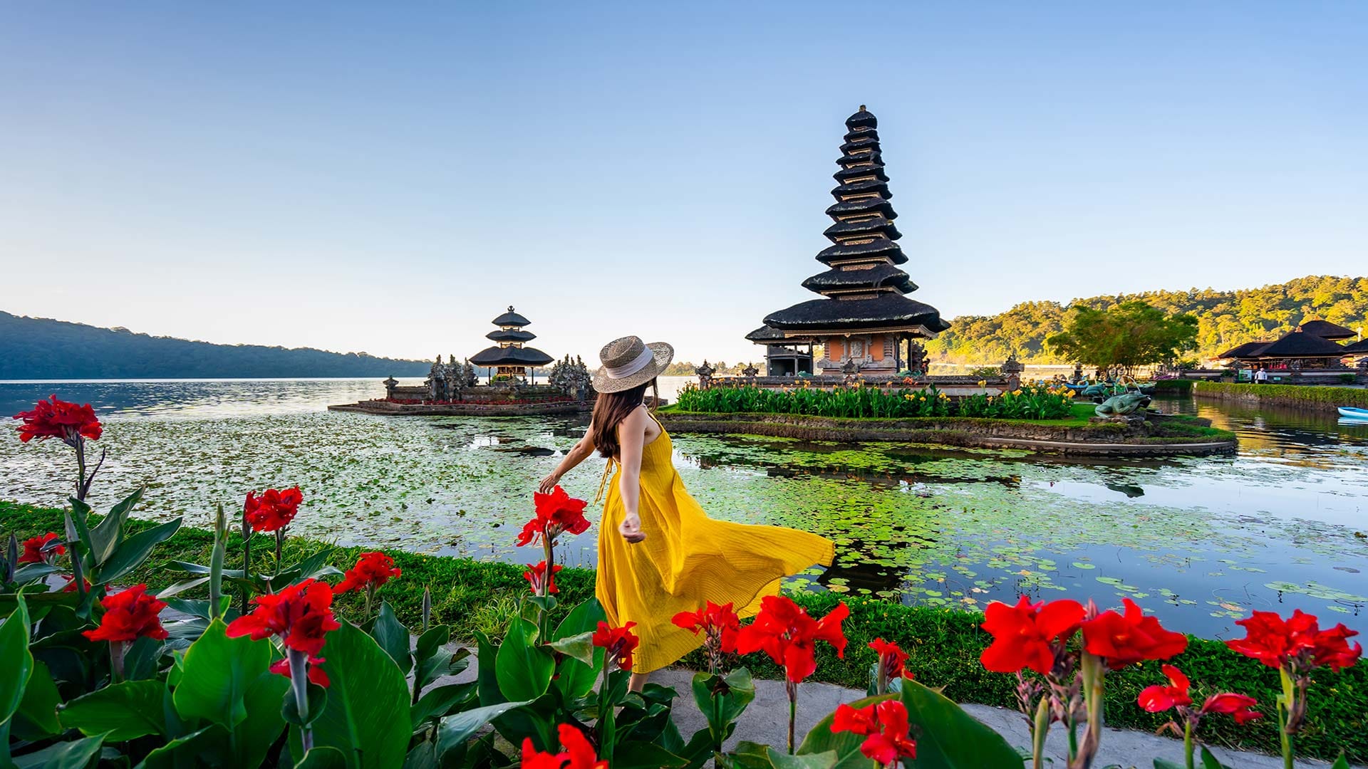 A vibrant scene of Ulun Danu Beratan Temple in Bali, with a woman in a flowing yellow dress surrounded by red flowers and serene lake waters.