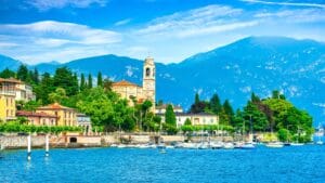 A scenic view of Como, Italy, showing colorful houses, a clock tower, and boats docked along the lake with mountains in the background.