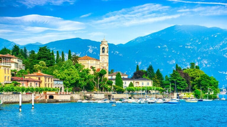 A scenic view of Como, Italy, showing colorful houses, a clock tower, and boats docked along the lake with mountains in the background.