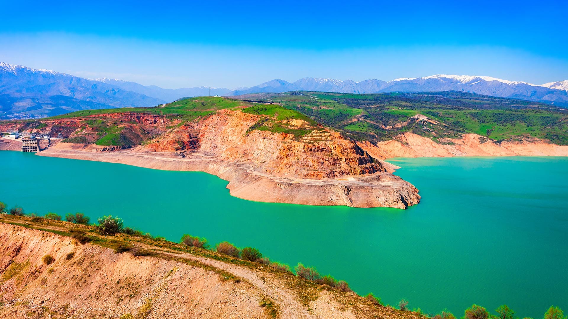 A vibrant view of the Charvak Reservoir with turquoise waters surrounded by lush green hills and rugged cliffs under a bright blue sky.