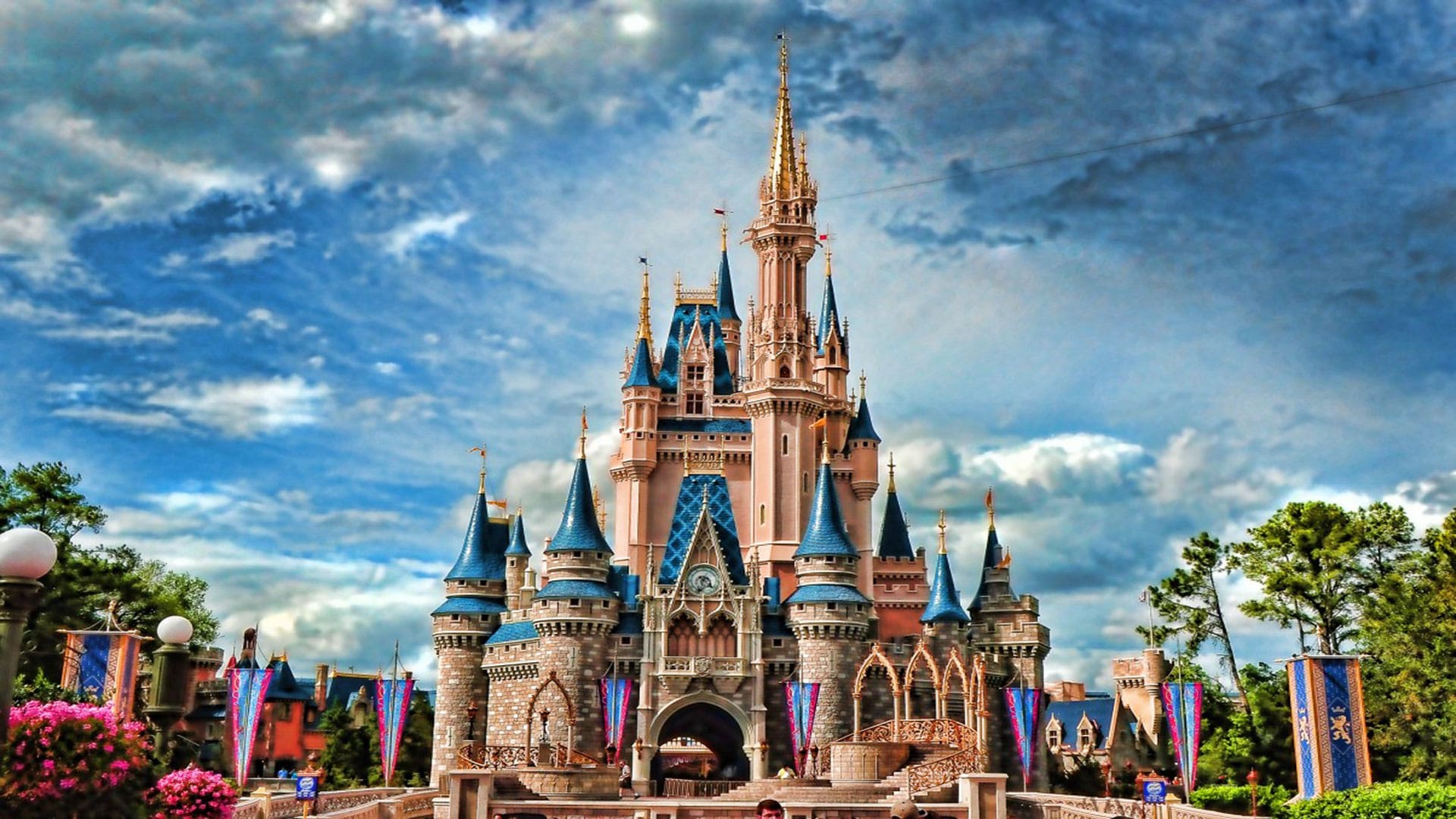 "Cinderella Castle at Disney World, with its enchanting spires against a dramatic sky, surrounded by colorful banners and lush greenery.