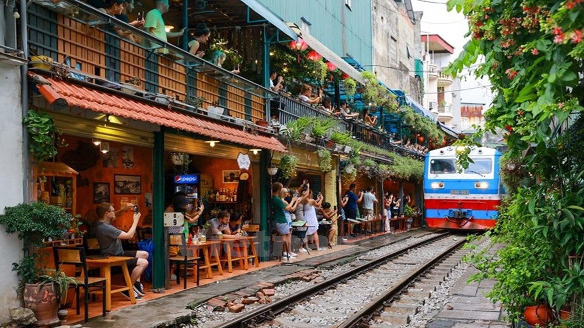 Train passing through Hanoi Train Street, surrounded by cafes and tourists taking photos.