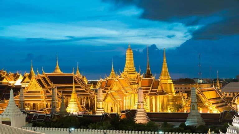 The illuminated spires and intricate rooftops of the Grand Palace in Bangkok against a dramatic evening sky.