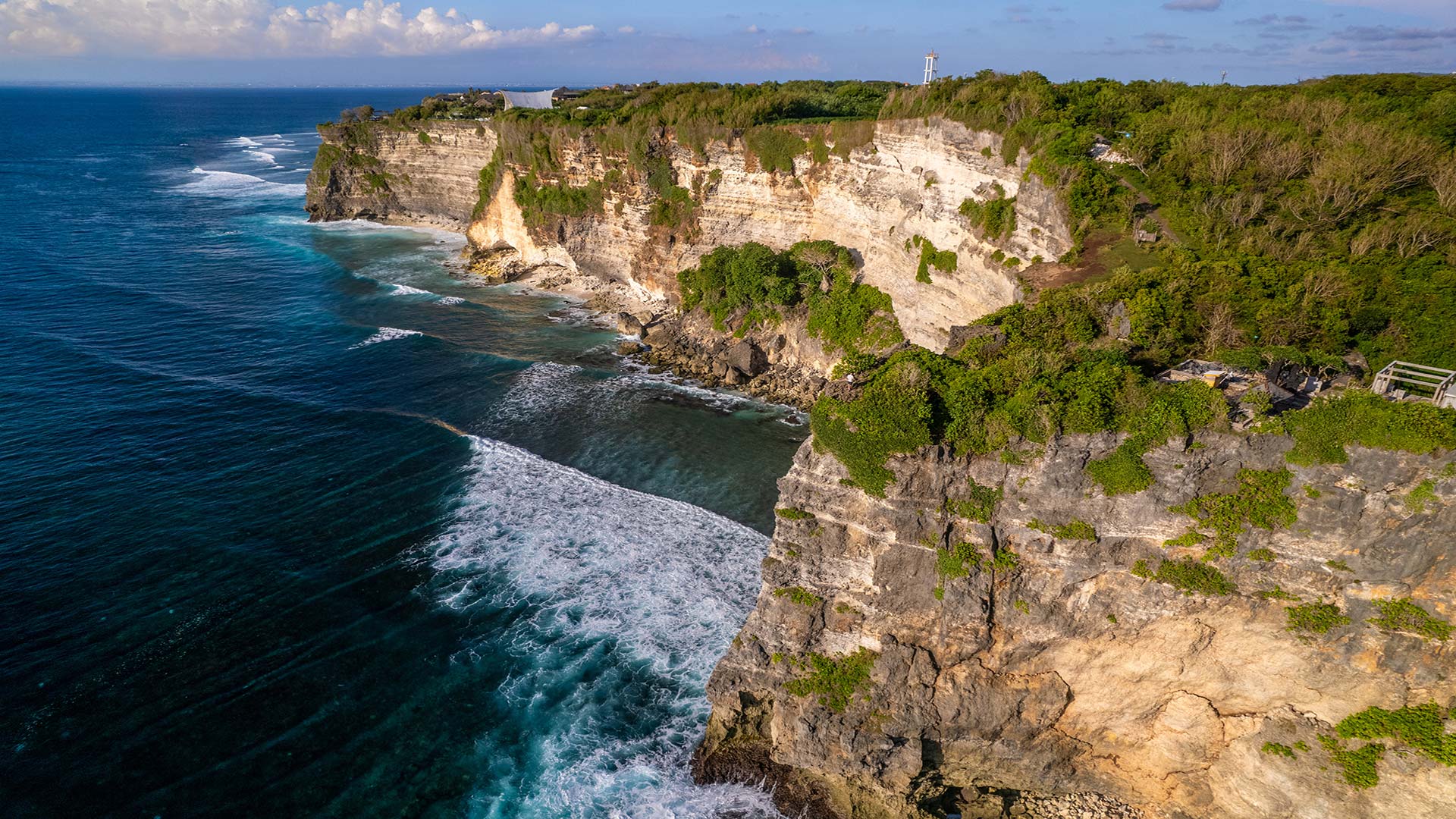 A stunning aerial view of the rugged cliffs of Uluwatu, bordered by the deep blue ocean waves crashing against the shoreline.