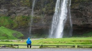 person in a blue raincoat sits on a bench admiring the majestic Seljalandsfoss waterfall in Iceland, surrounded by lush green scenery.
