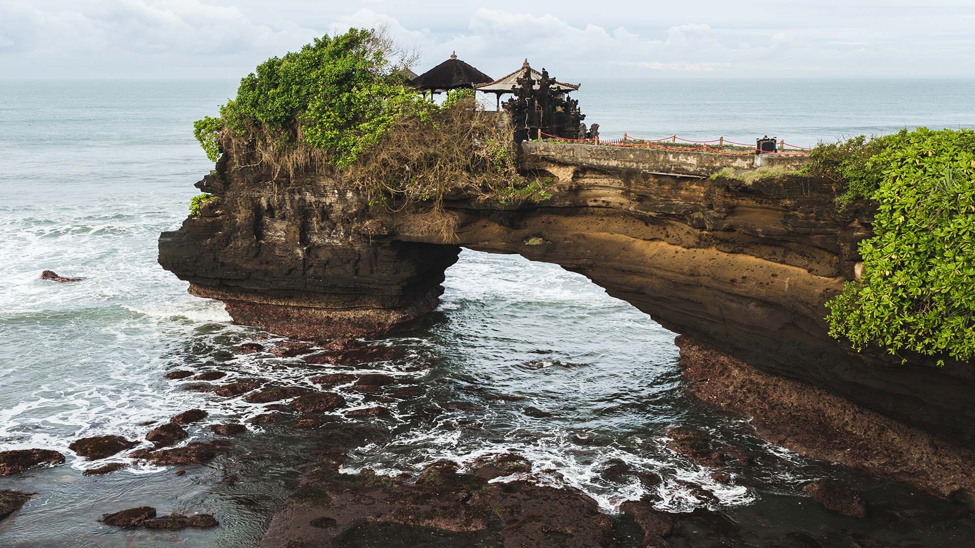 The iconic Tanah Lot Temple perched atop a natural rock formation, surrounded by the rolling waves of the Indian Ocean.