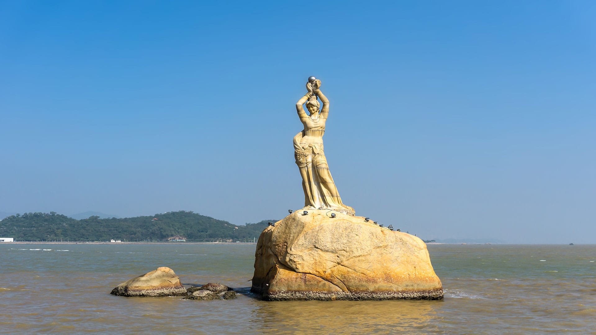 The iconic Fishing Girl Statue in Zhuhai, China, standing elegantly on a rock in the sea under a clear blue sky.